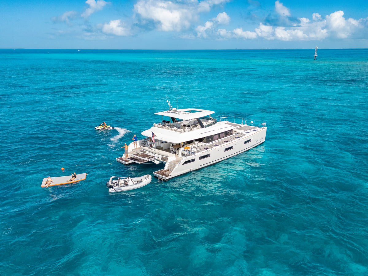 A white luxury catamaran yacht floating on bright turquoise ocean water under a blue sky with white clouds, with small boats nearby.