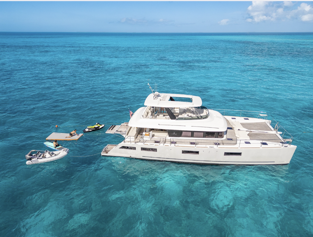 A white luxury catamaran yacht floating on clear turquoise ocean water, with small boats and a dock nearby under a blue sky.