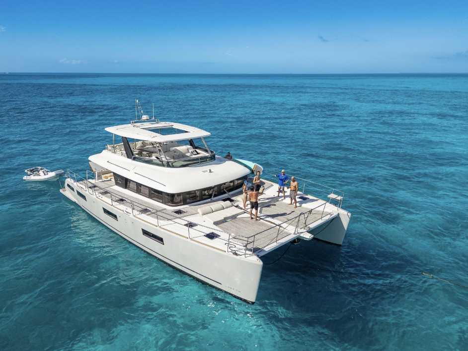 A white luxury catamaran yacht floating on clear turquoise ocean water under a blue sky, with several people standing on the deck and a small boat nearby.