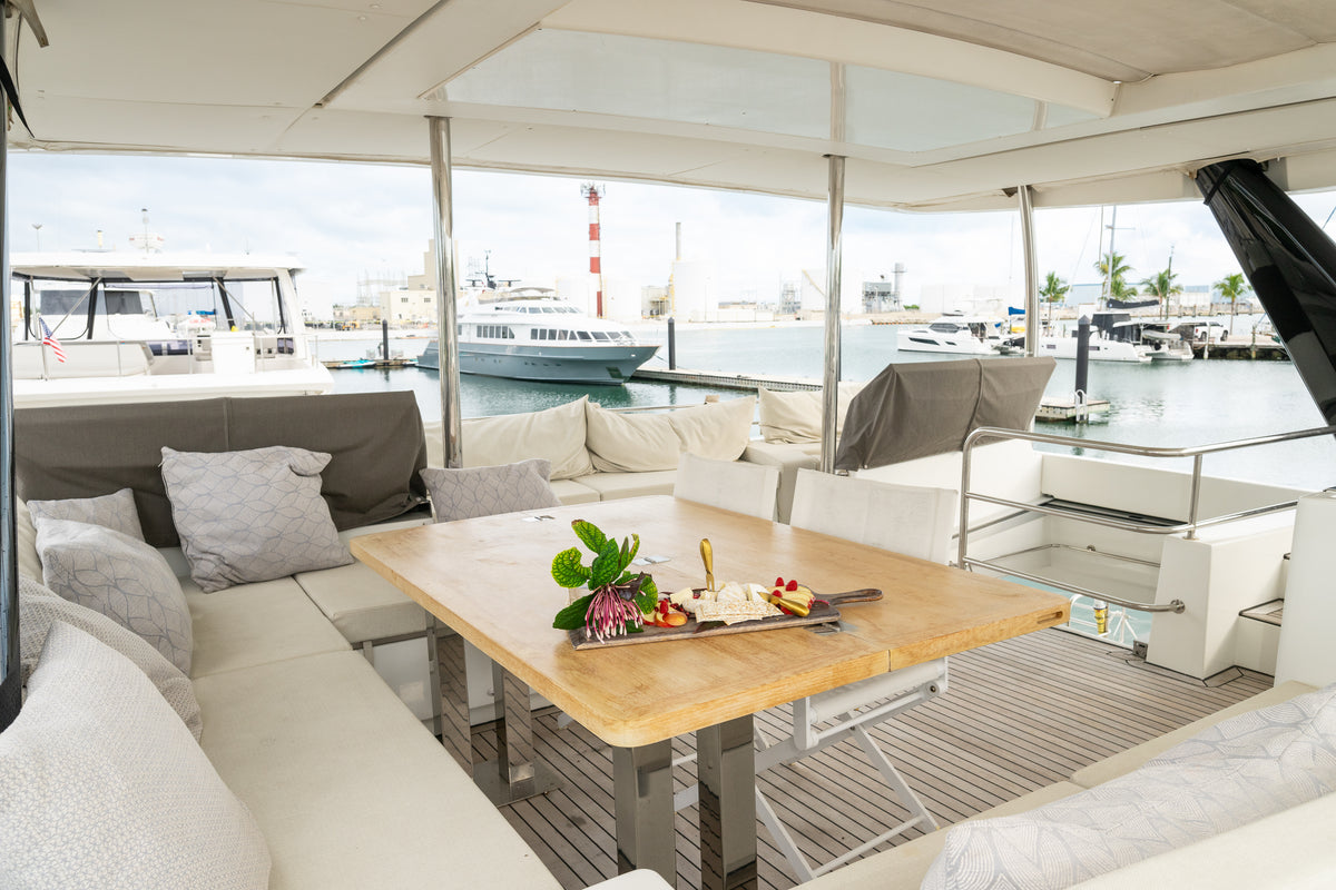 Outdoor dining area on a yacht or boat with a light wood table, cream upholstered seating, gray pillows, and large windows overlooking a marina.