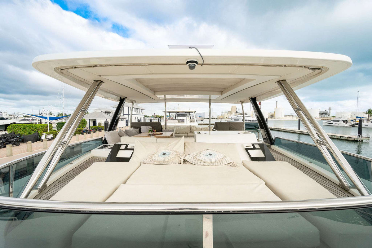 A spacious outdoor sun pad area on the deck of a motor yacht, featuring cream-colored cushions under a white hardtop canopy, with a marina and boats visible in the background under a cloudy sky.