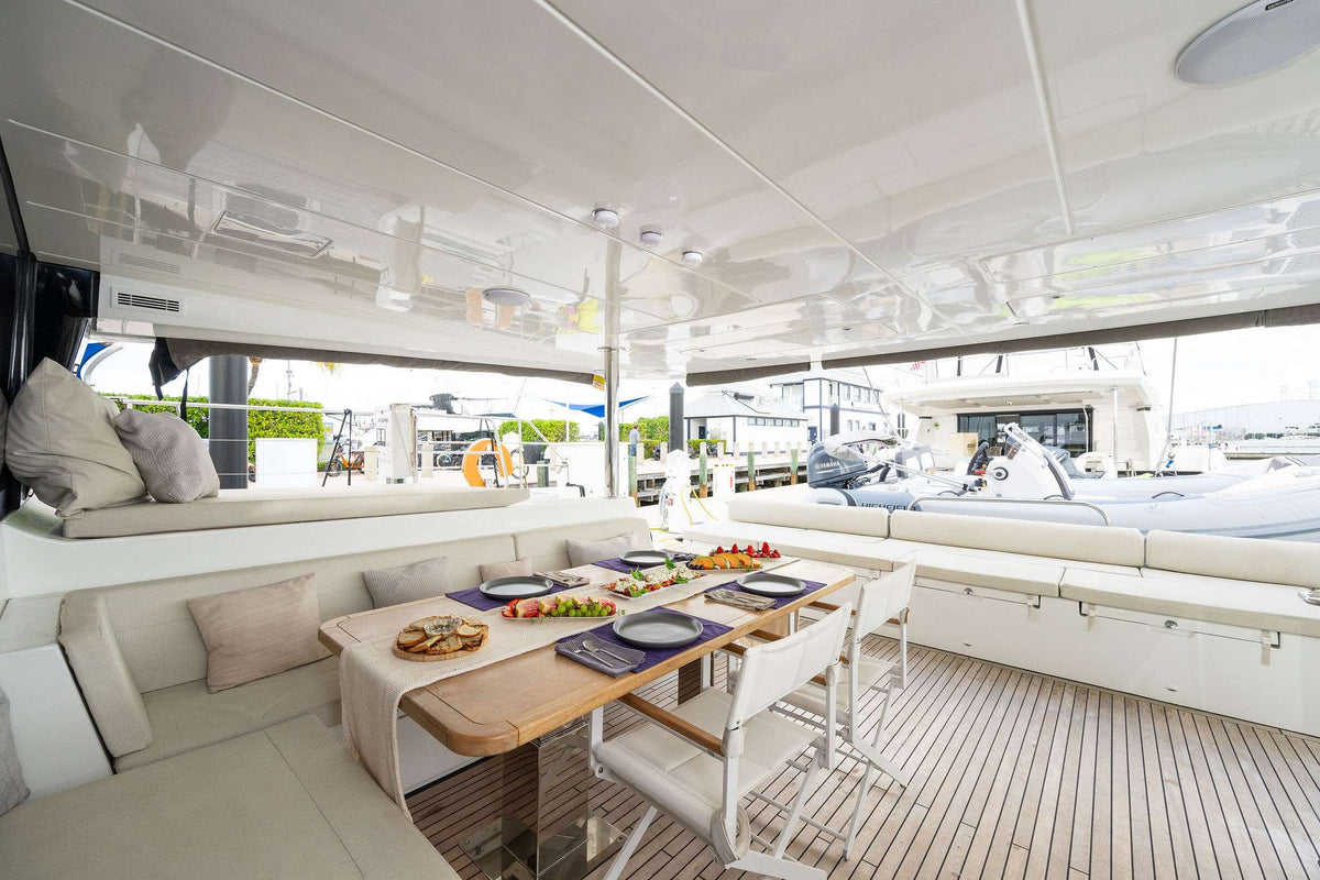 The interior of a luxury yacht featuring a dining area with a wooden table, white chairs, and a sunken seating area with cushions, set against a white ceiling and a wooden deck.