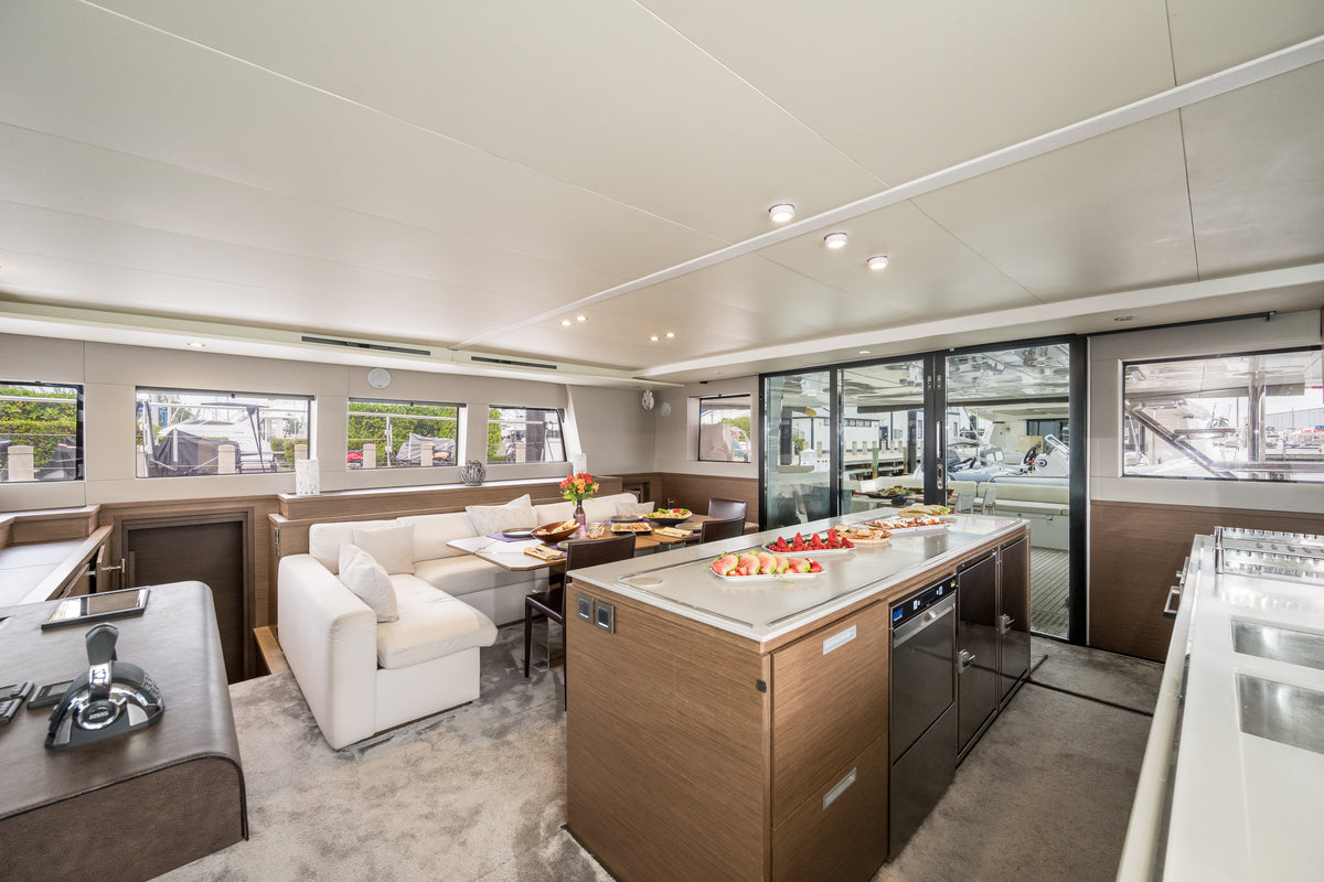 A luxury yacht interior featuring a modern galley kitchen with a wood-look island, white countertops, and stainless steel appliances, alongside a white lounge chair and dining area with large windows.