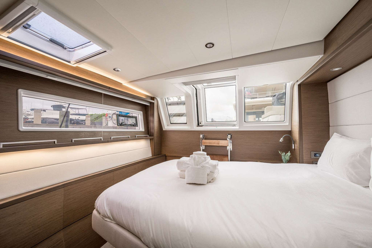 A modern yacht or cruise ship cabin featuring a large bed with white linens, wood-paneled walls, skylights, and windows with a view of the deck outside.