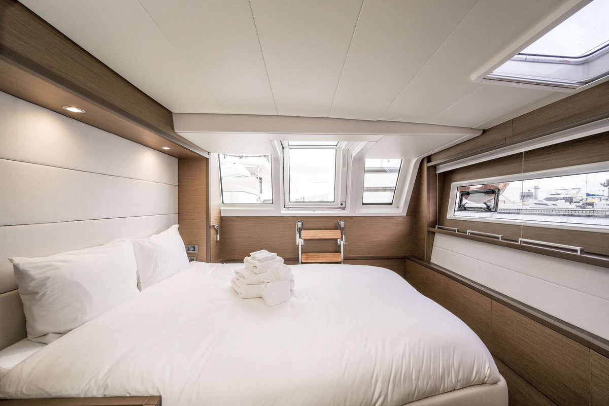 A modern yacht or boat cabin featuring a large bed with white bedding, a folded towel, wood paneling, and multiple windows with skylights.