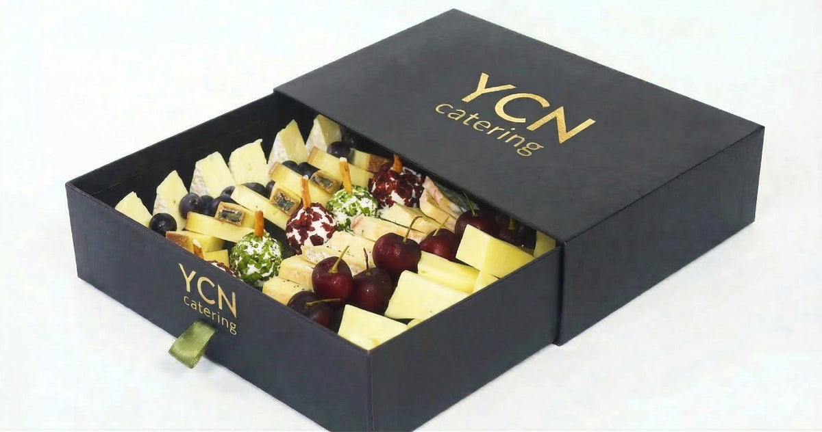 A YCN catering cheese and charcuterie gift box, shown open to reveal an assortment of cheeses, grapes, and cured meats inside a black box with gold lettering.