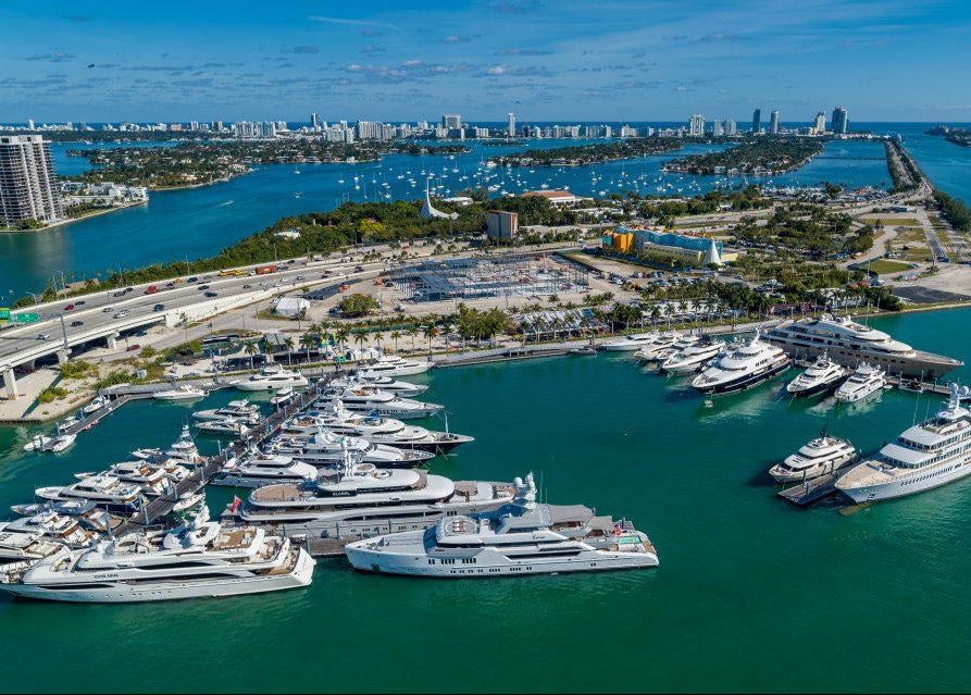 Aerial view of a marina filled with white yachts and boats, surrounded by turquoise water, with a city skyline and bridge in the background under a blue sky.