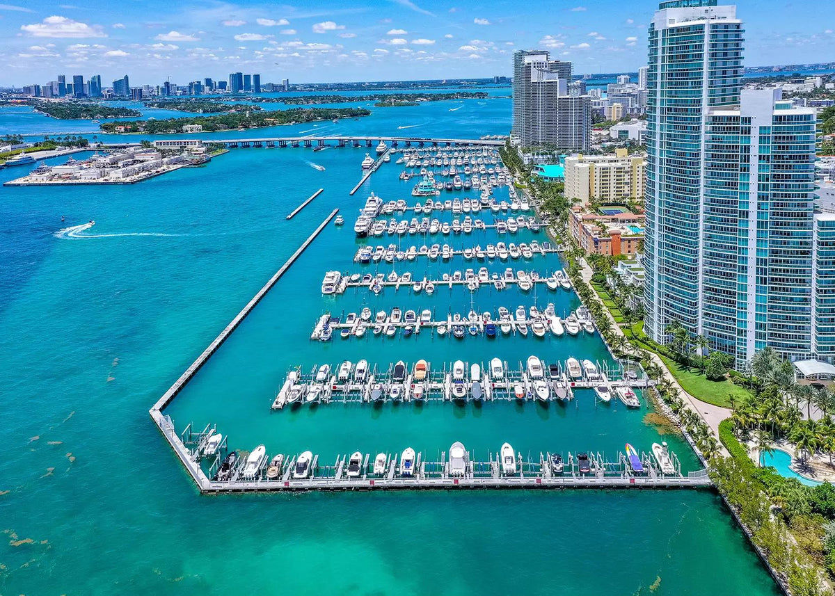 Aerial view of a marina filled with boats, bordered by tall modern high-rises, turquoise water, and a distant city skyline under a blue sky.