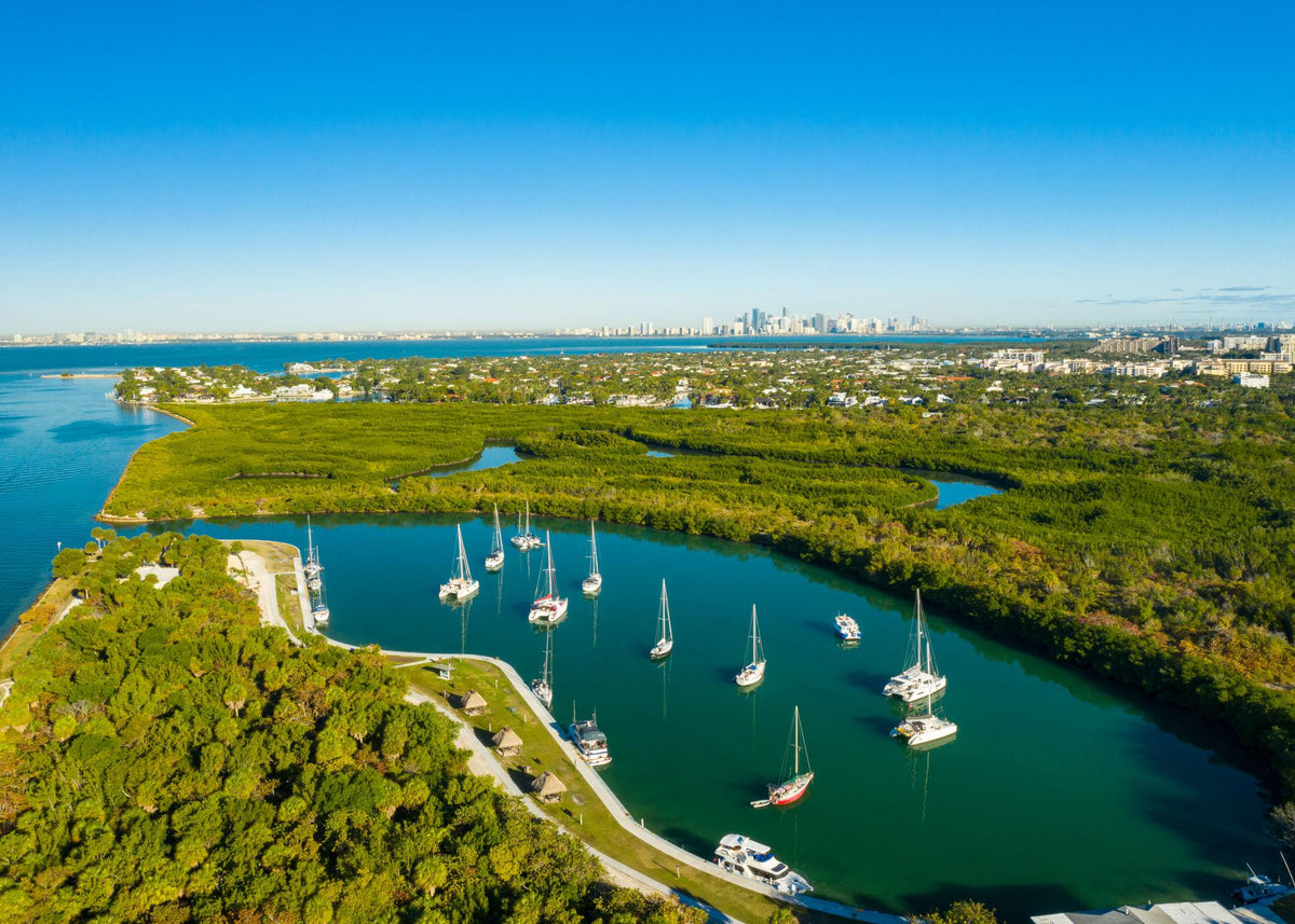 Aerial view of a tropical waterway with sailboats on turquoise water, surrounded by dense green mangroves and leading toward a distant city skyline under a clear blue sky.