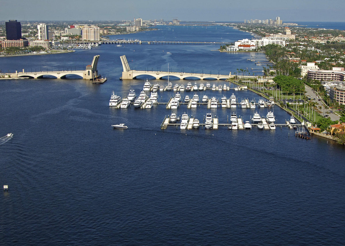 Aerial view of a marina filled with white boats and yachts, bordered by waterfront buildings and green trees, with a drawbridge spanning deep blue water in the background.