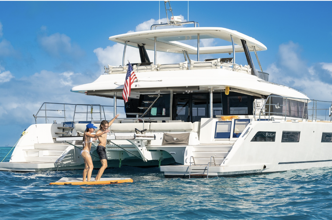 A large white catamaran yacht floating on blue ocean water under a bright blue sky with clouds. Two people are standing on a yellow board at the swim platform on the front of the yacht, with an American flag hanging near the cockpit.