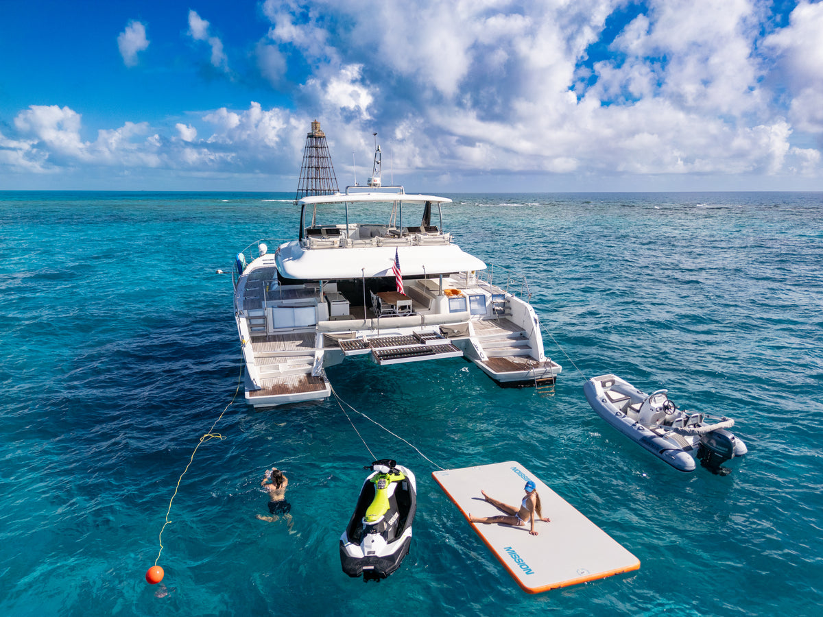 A luxury white catamaran floating in turquoise ocean water under a blue sky with white clouds, with a jet ski, inflatable paddle board, and small rigid inflatable boat nearby, and a person lying on the paddle board.
