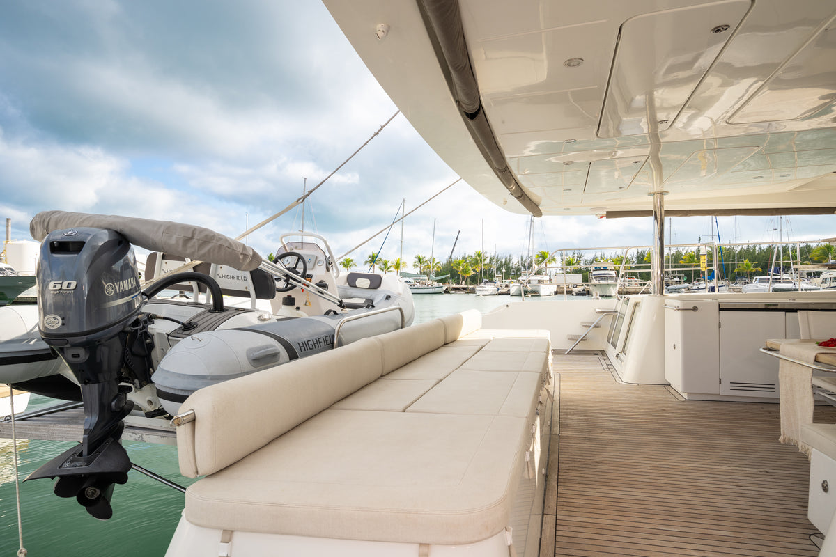 A luxury yacht deck featuring a cream-colored cushioned lounge bench, a wooden walkway, and a Yamaha outboard motor attached to a rigid inflatable boat (RIB) docked alongside under a cloudy sky.