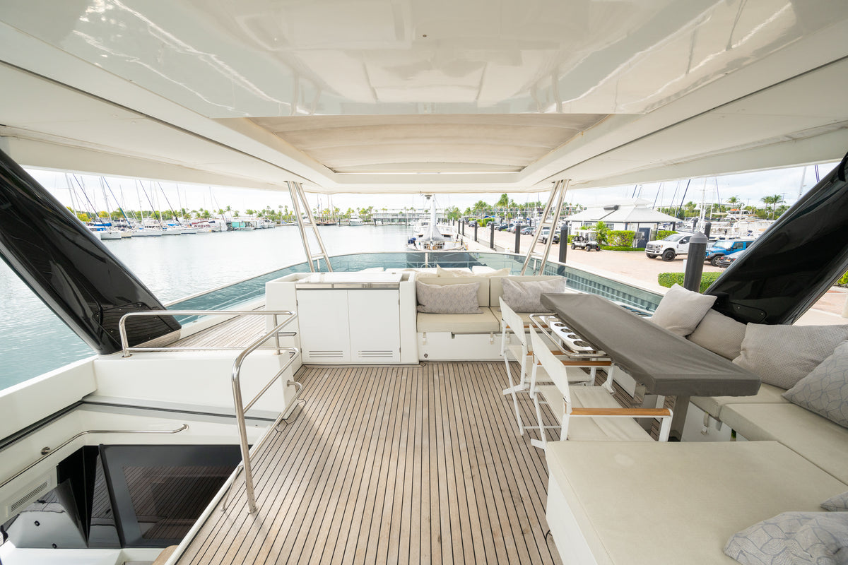 The upper deck of a luxury yacht featuring a teak wood flooring, a white hardtop canopy, a rear seating area with beige cushions, and an outdoor dining set with a gray table, overlooking a marina background.