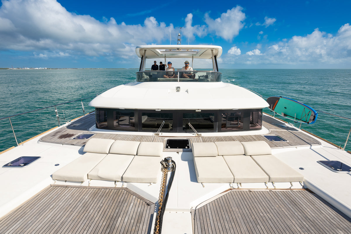 A luxury yacht deck viewed from the stern, featuring cream-colored cushioned lounge chairs, a wooden deck, a green and blue surfboard, and an open ocean backdrop under a blue sky with white clouds.