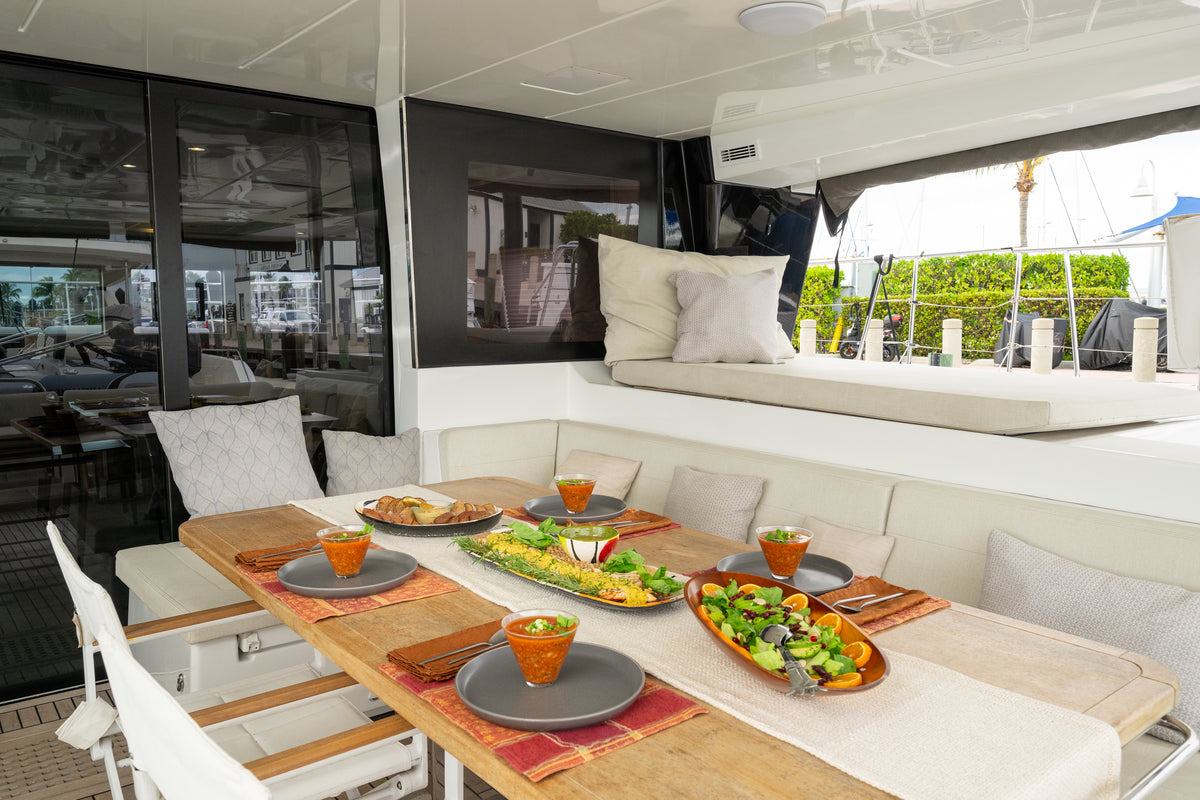 A modern yacht dining area with a wooden table set for six, featuring gray plates, orange drink glasses, a salad platter, and a main dish, surrounded by cream upholstered chairs and a daybed with pillows.