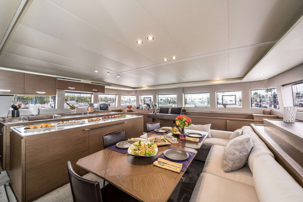 A spacious yacht or boat interior dining and galley area with wood cabinetry, cream seating, large windows, and a dining table set with plates and a salad.