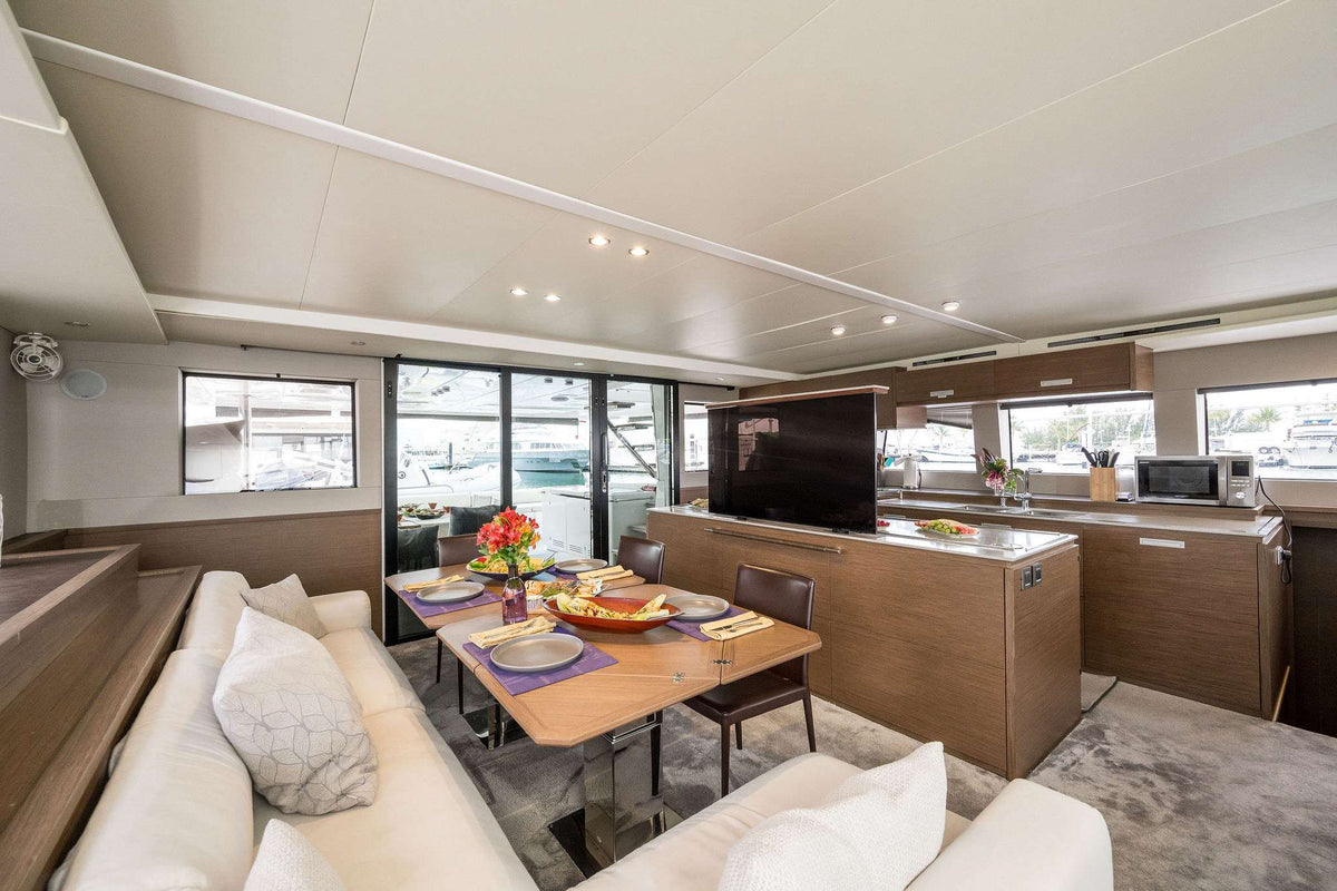 The interior of a modern yacht or houseboat featuring a dining area with a wooden table and chairs, a cream-colored sofa with pillows, and a galley kitchen with wood cabinetry and a black television mounted on the wall.