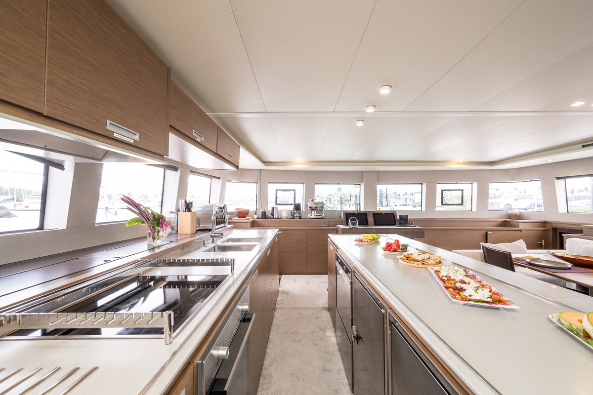 A modern yacht galley kitchen featuring light wood cabinetry, stainless steel appliances, and a long central island with plated food, set against large windows.