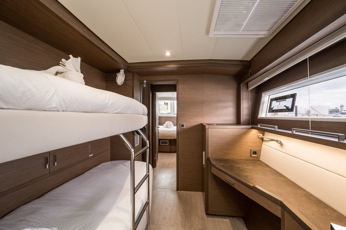 A modern boat cabin interior featuring a double bunk bed with white bedding, a wooden desk and vanity, and a large window looking out onto water.