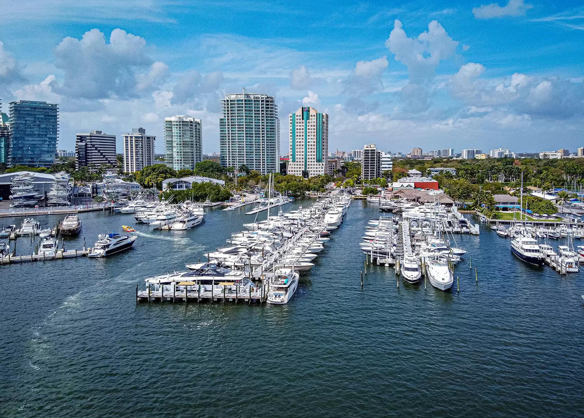 Aerial view of a marina filled with white yachts and boats, with a city skyline of modern high-rise buildings under a bright blue sky with scattered clouds.