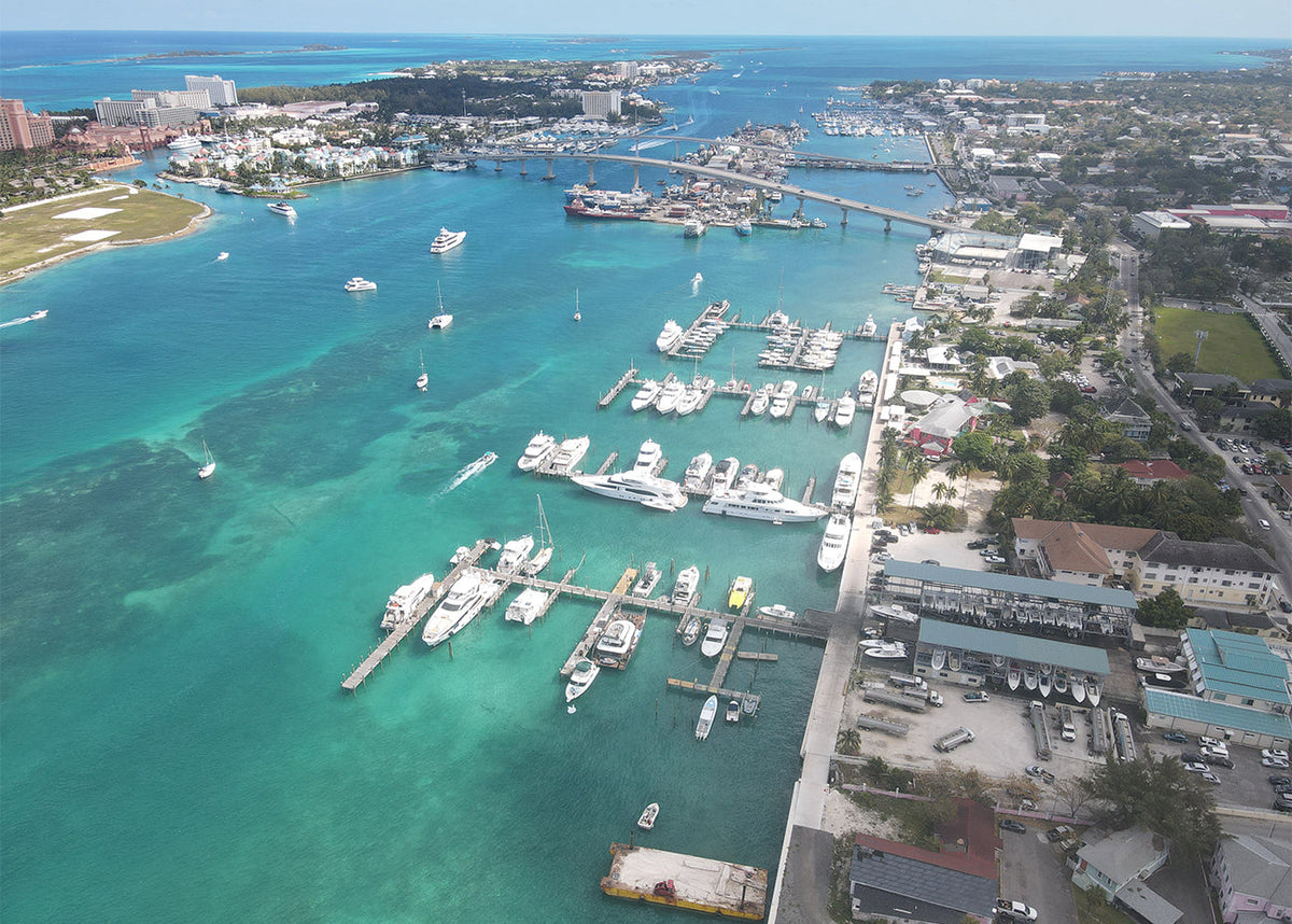 Aerial view of a marina with numerous white boats and yachts docked in clear turquoise water, with a shoreline, bridges, and buildings visible in the background.