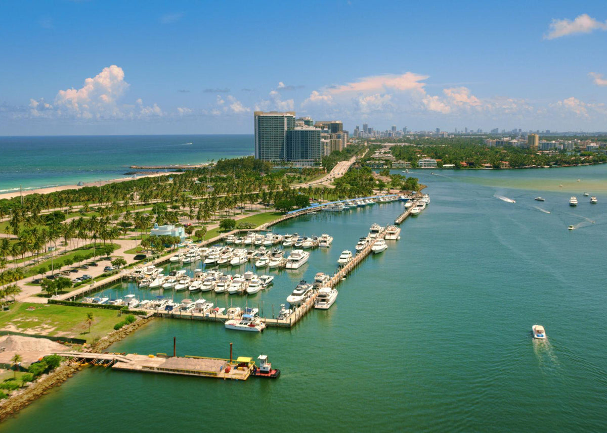 Aerial view of a tropical coastal marina with numerous white boats docked in turquoise water, palm trees, beachfront properties, and a city skyline under a blue sky with scattered clouds.
