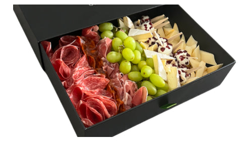 A black rectangular gift box filled with an assorted charcuterie and cheese spread, including sliced cured meats, green grapes, and wedges of cheese, photographed on a white background.