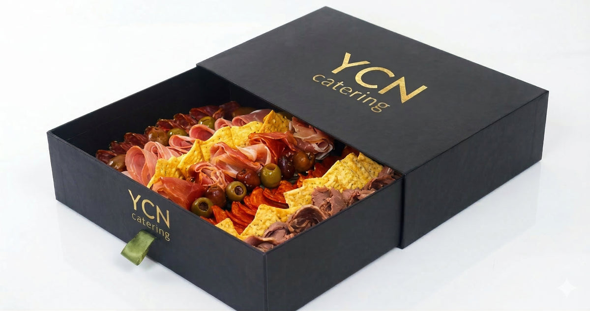A black YCN catering gift box, partially open to reveal a charcuterie board filled with crackers, salami, prosciutto, cheese, olives, and other appetizers, set against a clean white background.