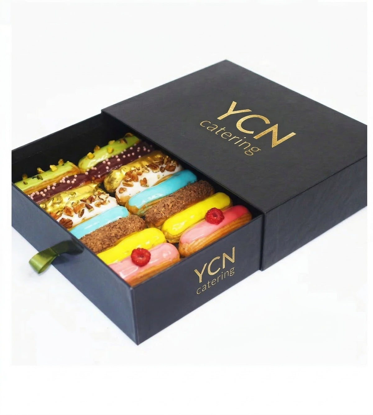 A black YCN catering gift box with the lid partially open, revealing assorted colorful pastries and croissants inside. The box lid and exterior are black with gold YCN catering branding.
