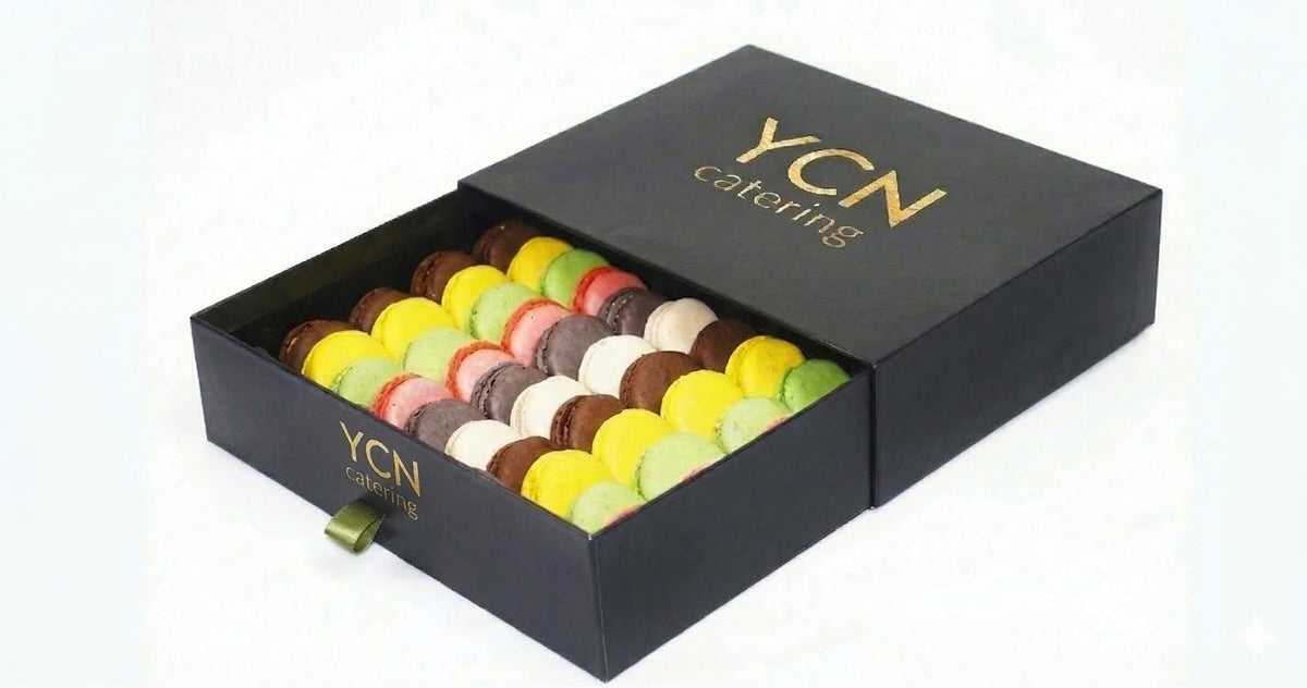A black YCN Catering gift box filled with assorted colorful macarons, shown on a clean white background.