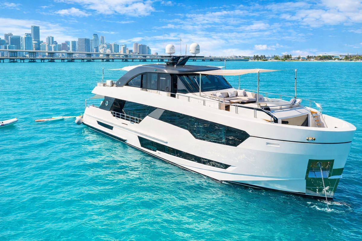 A luxury motor yacht with a white hull and dark glass superstructure floating on bright turquoise water, with a city skyline and bridge visible in the background under a blue sky.