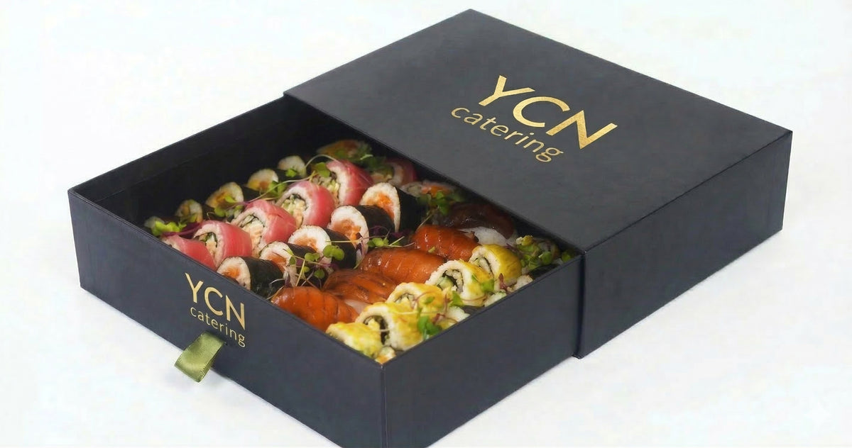 A black YCN catering gift box with the lid propped open, revealing a colorful assortment of sushi rolls inside. The box features gold foil branding on both the lid and the front panel, set against a plain white background.