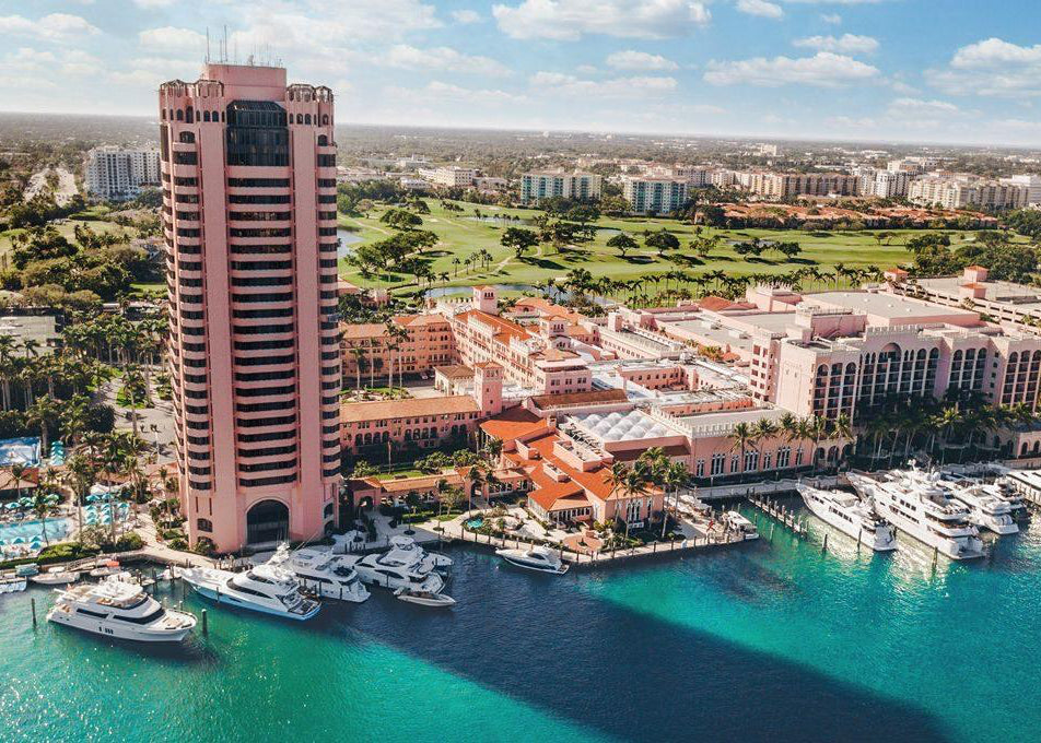 Aerial view of a large pink resort hotel with a tall tower, marina with many white yachts, turquoise water, golf land, and distant city buildings under a blue sky.